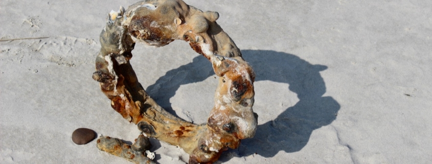 Auf hellem Sand steht ein Ring aus Metall, verkrustet, aufgequollen und voller Beulen aus Rost, wahrscheinlich ein altes Bullauge, das sehr lange im Meer unterwegs war. Foto: Birte Vogel