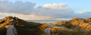 Zwei Wege in den Dünen: ein Bohlenweg links, ein Sandweg in der Mitte, beide führen runter zum Strand und zum Meer, die man im Hintergrund sieht. Darüber einige Wolken, eine regnet sich über dem Meer ab, und ganz oben hellblauer Himmel. Foto: Birte Vogel