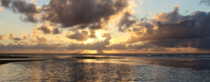 Sonnenuntergang am Meer: Die Priele sind vollgelaufen, nur ein bisschen Sand schaut noch raus, über dem Wasser Wolken, hinter denen die Sonne untergeht. Hinter einer längeren Wolke sieht man die Sonnenstrahlen hindurchkommen. Foto: Birte Vogel