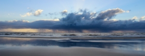 Blick vom Strand aufs Meer, über dem eine breite Regenwolke hängt und sich abregnet. Sie spiegelt sich im Wasser auf dem Sand. Darüber hellblauer Himmel. (Foto: Birte Vogel)