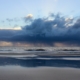 Blick vom Strand aufs Meer, über dem eine breite Regenwolke hängt und sich abregnet. Sie spiegelt sich im Wasser auf dem Sand. Darüber hellblauer Himmel. (Foto: Birte Vogel)