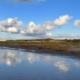 Eine Reihe von Dünen schieben sich von rechts bis nach links durchs Bild, teilweise noch grün, teilweise schon orangegelb. Im Vordergrund steht Wasser, über den Dünen blauer Himmel mit einigen Wolken, die sich unten im Wasser spiegeln. (Foto: Birte Vogel)