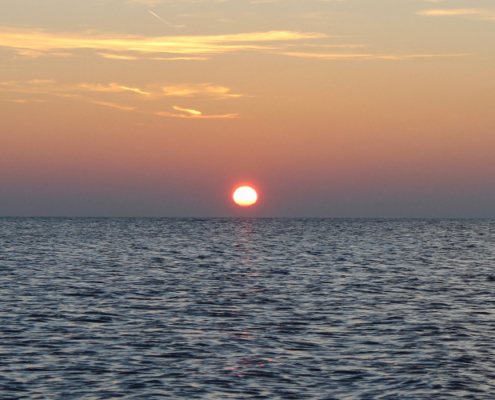 Sonnenuntergang am Meer, im Vordergrund ein leicht gewelltes, dunkelblaues Meer, darüber ein rötlich gelber Himmel mit wenigen goldfarbenen Wolkenschleiern, und in der Bildmitte die Sonne als rote Kugel. Foto: Birte Vogel