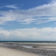 Im Vordergrund ein menschenleerer Strand, durchzogen von einem kleinen Pril, im Hintergrund dunkelblau das Meer, darüber ein blauer Himmel mit vielen weißen Wolken. Foto: Birte Vogel