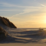 Links im Vordergrund Dünen, rechts erstreckt sich der Strand, am Horizont das Meer darüber eine gleißende Sonne kurz vorm Sonnenuntergang, noch hellgoldfarben. Foto: Birte Vogel