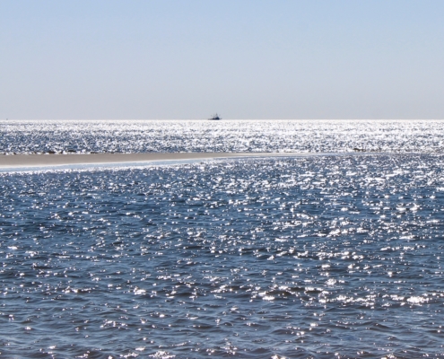 Ein ganz blaues Meer im Vordergrund, das zwar unruhig ist, aber nur kleine Wellenbewegungen zeigt. Von links kommt mittig eine kleine Sandbank ins Bild. Am Horizont ein Krabbenkutter. Der Himmel darüber hellblau. (Foto: Birte Vogel)
