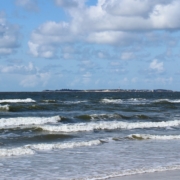 Wellen auf einer leuchtend dunkelblau-grünen Nordsee. Am Horizont die Insel Sylt. Darüber ein blauer Himmel mit einigen Schäfchenwolken. (Foto: Birte Vogel)