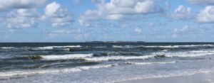 Wellen auf einer leuchtend dunkelblau-grünen Nordsee. Am Horizont die Insel Sylt. Darüber ein blauer Himmel mit einigen Schäfchenwolken. (Foto: Birte Vogel)
