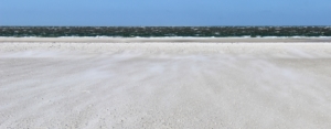 Weißer Sandstrand, über den der Sand vom dunkelgrünen, unruhigen Meer auf dich zugeweht wird. Darüber ein leuchtend blauer Himmel. (Foto: Birte Vogel)