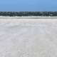 Weißer Sandstrand, über den der Sand vom dunkelgrünen, unruhigen Meer auf dich zugeweht wird. Darüber ein leuchtend blauer Himmel. (Foto: Birte Vogel)