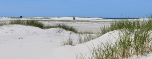 Ein Blick durch niedrige Sanddünen mit weißem Sand und einigen grünen Dünengräsern, über eine weite Sandfläche, am Horizont weitere niedrige Dünen, davor ein weiß-blau gestreifter Strandkorb. Im Hintergrund das blaue Meer, darüber ein blauer Himmel. (Foto: Birte Vogel)