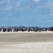 Im Vordergrund weißer Strand, in der Mitte eine große Anzahl bunter Strandkörbe, die in alle möglichen Richtungen stehen, am Horizont die Insel Sylt, darüber ein blauer Himmel mit einigen Wolken (Foto: Birte Vogel)