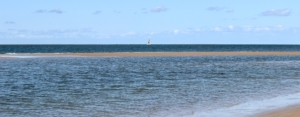 Im Vordergrund ein dunkelblaues Meer, in der Mitte, einmal quer von rechts nach links, eine noch feuchte Sandbank, dahinter am Horizont ein einsames Segelboot mit weiß-orangefarbenem Segel. Der Himmel ist hellblau mit wenigen kleinen Wolken. (Foto: Birte Vogel)